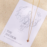 Gold necklace with small triangle red garnet pendant on a affirmation card with 'The Anchor' design and text.