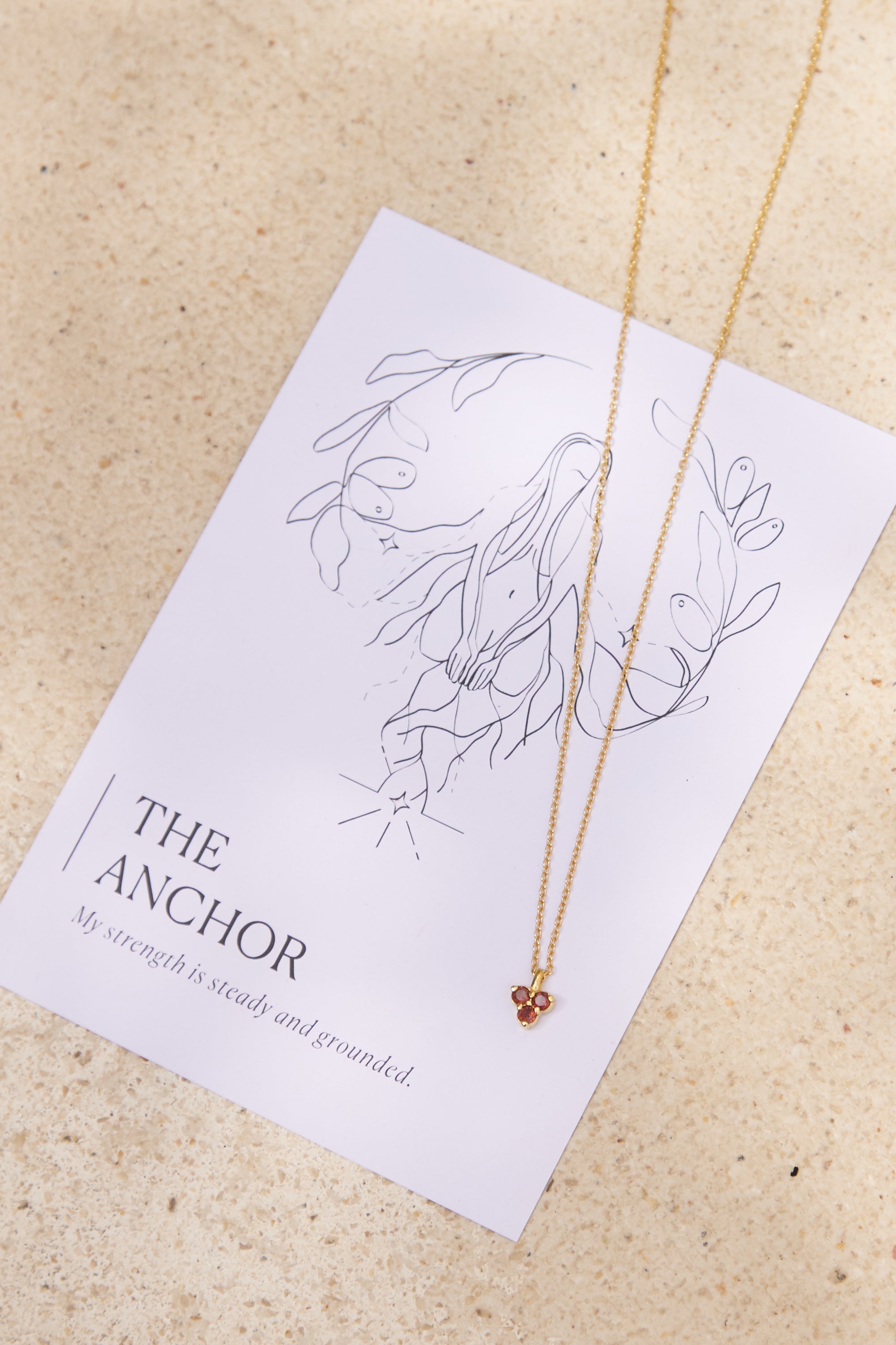 Gold necklace with small triangle red garnet pendant on a affirmation card with 'The Anchor' design and text.