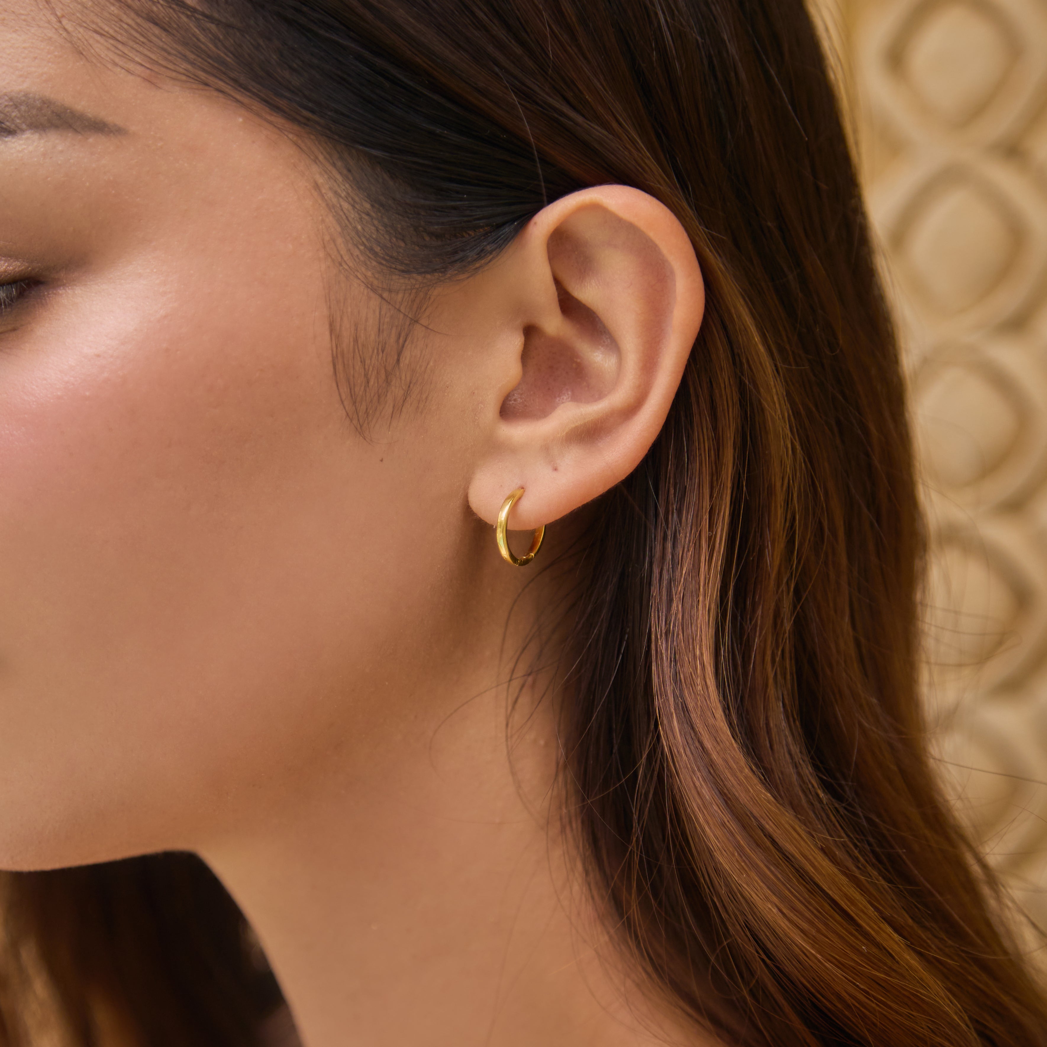 Close-up of a person wearing a small gold hoop earring with a textured beige wall background