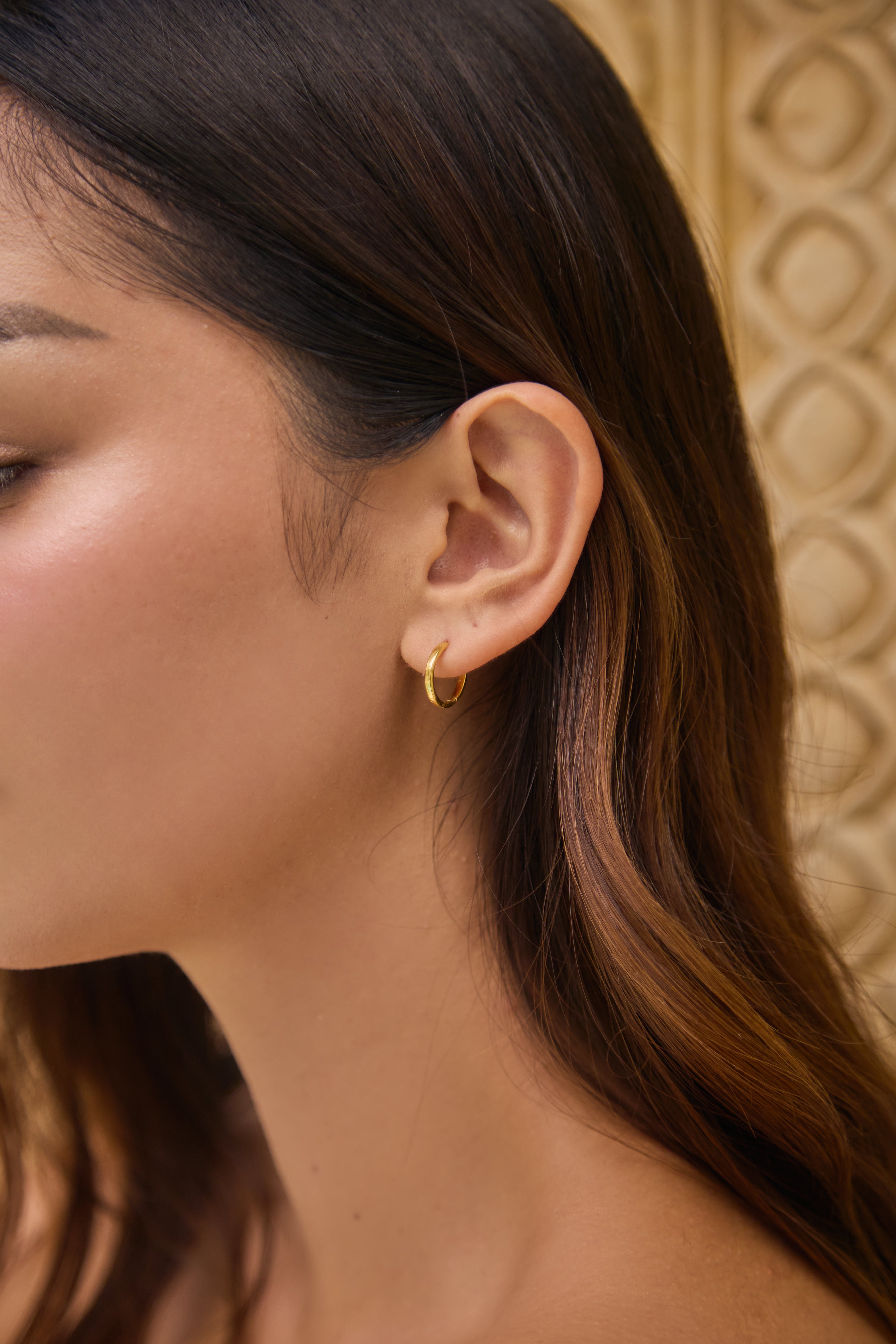 Close-up of a person wearing a small gold hoop earring with a textured beige wall background