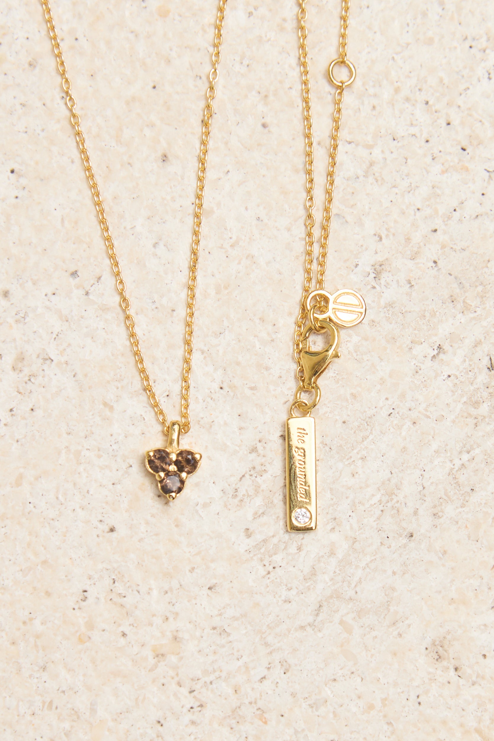 Gold necklace with triangle smoky quartz pendant and bar pendant on a beige background