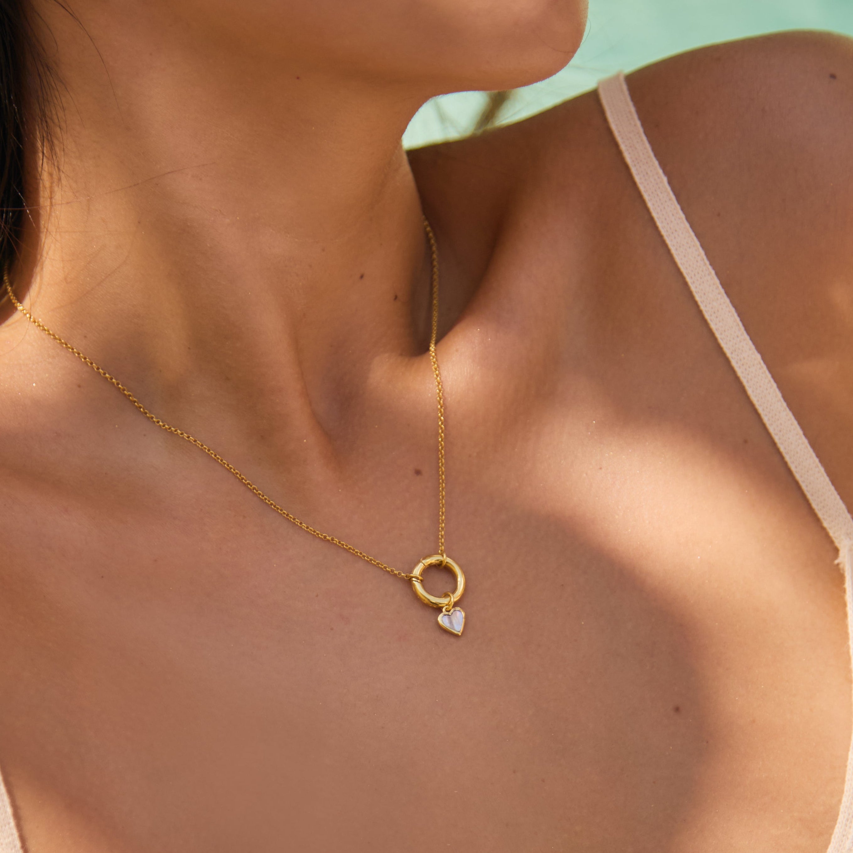Close-up of a person wearing a gold necklace with a heart pendant against a blurred natural background.