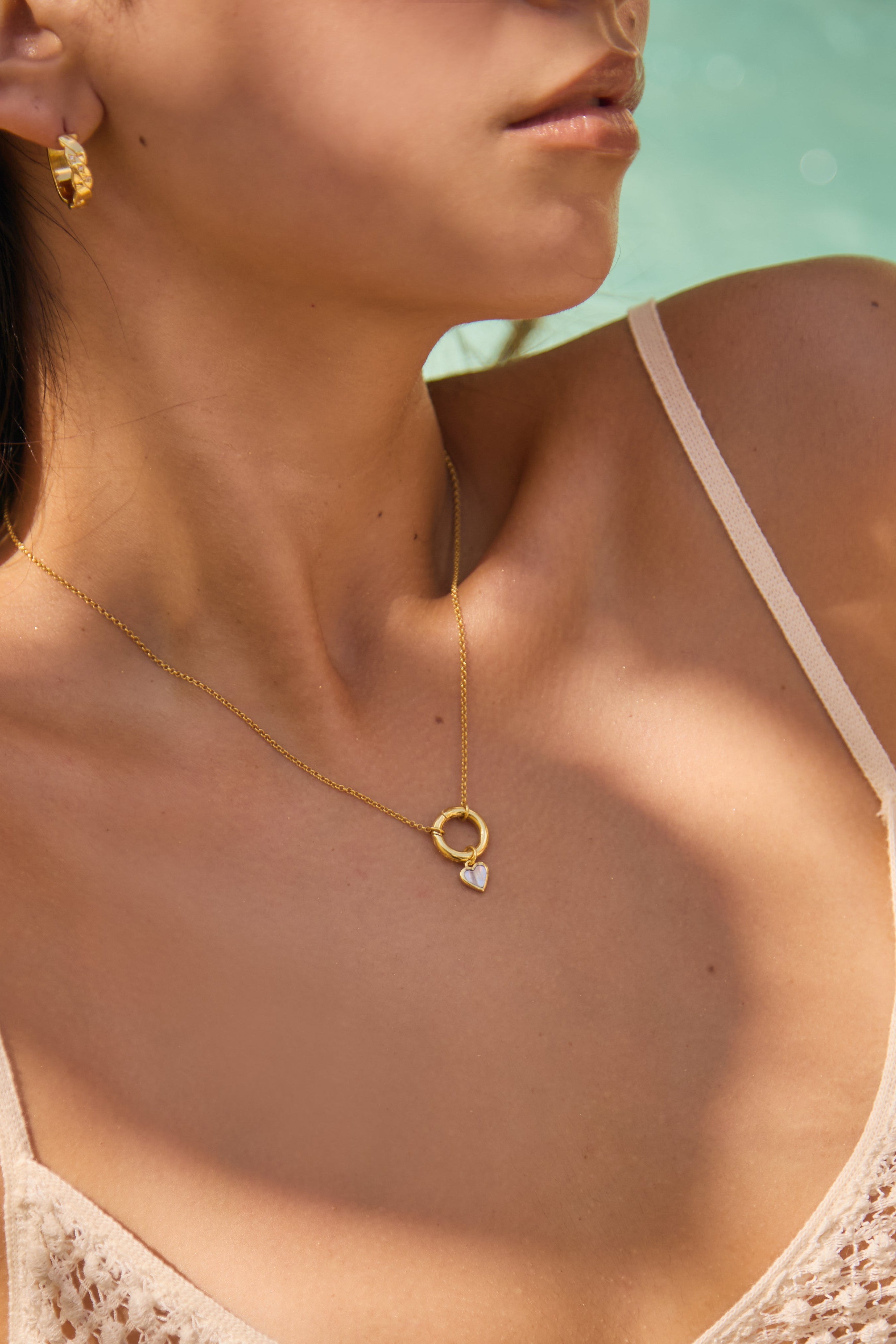 Close-up of a person wearing a gold necklace with a heart pendant against a blurred natural background.