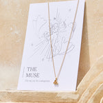 Gold necklace with a yellow citrine triangle pendant on a 'The Muse' affirmation card on a beige background