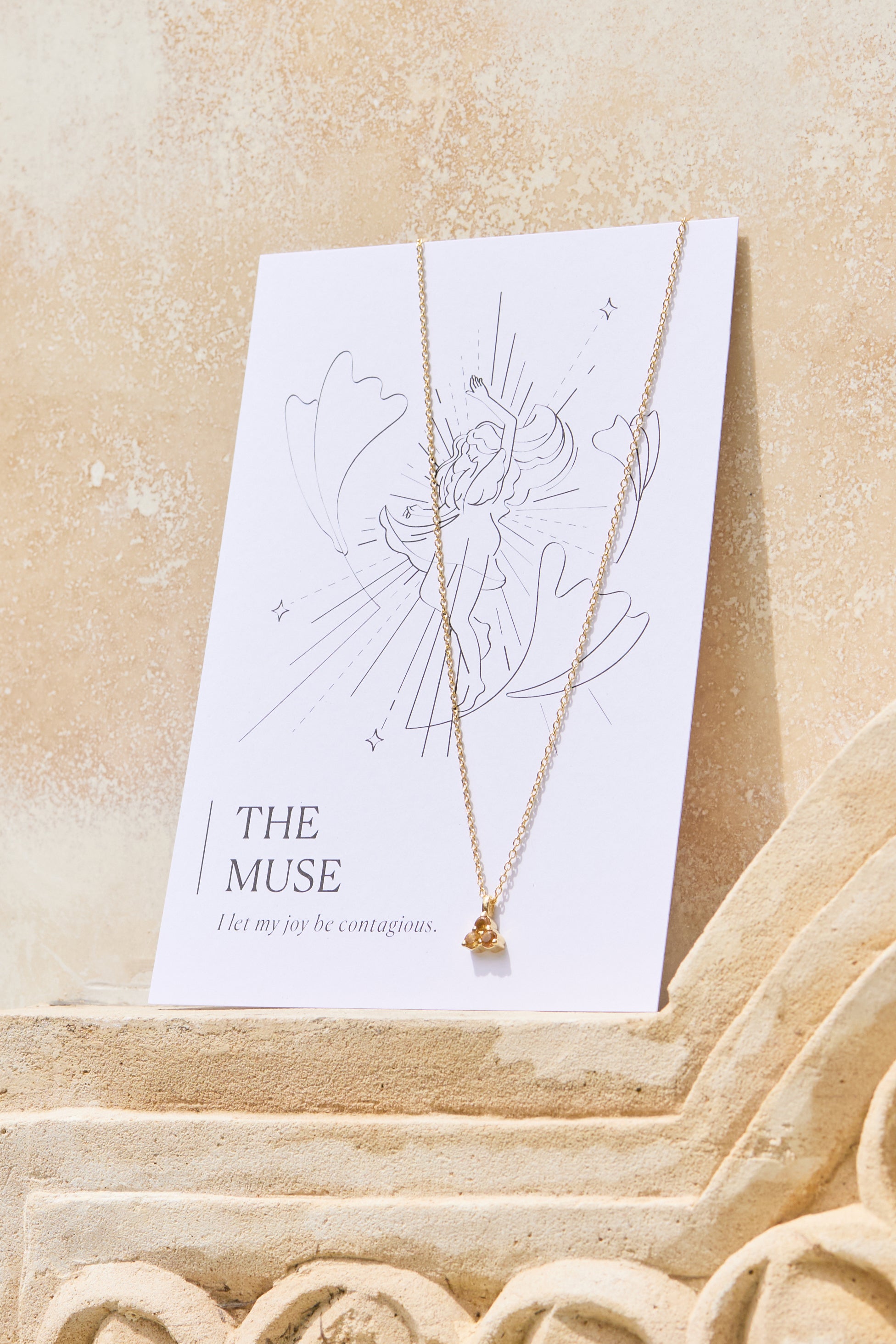 Gold necklace with a yellow citrine triangle pendant on a 'The Muse' affirmation card on a beige background
