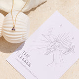 Necklace with a small triangle blue iolite pendant on a affirmation card titled 'The Seeker' on a sandy background