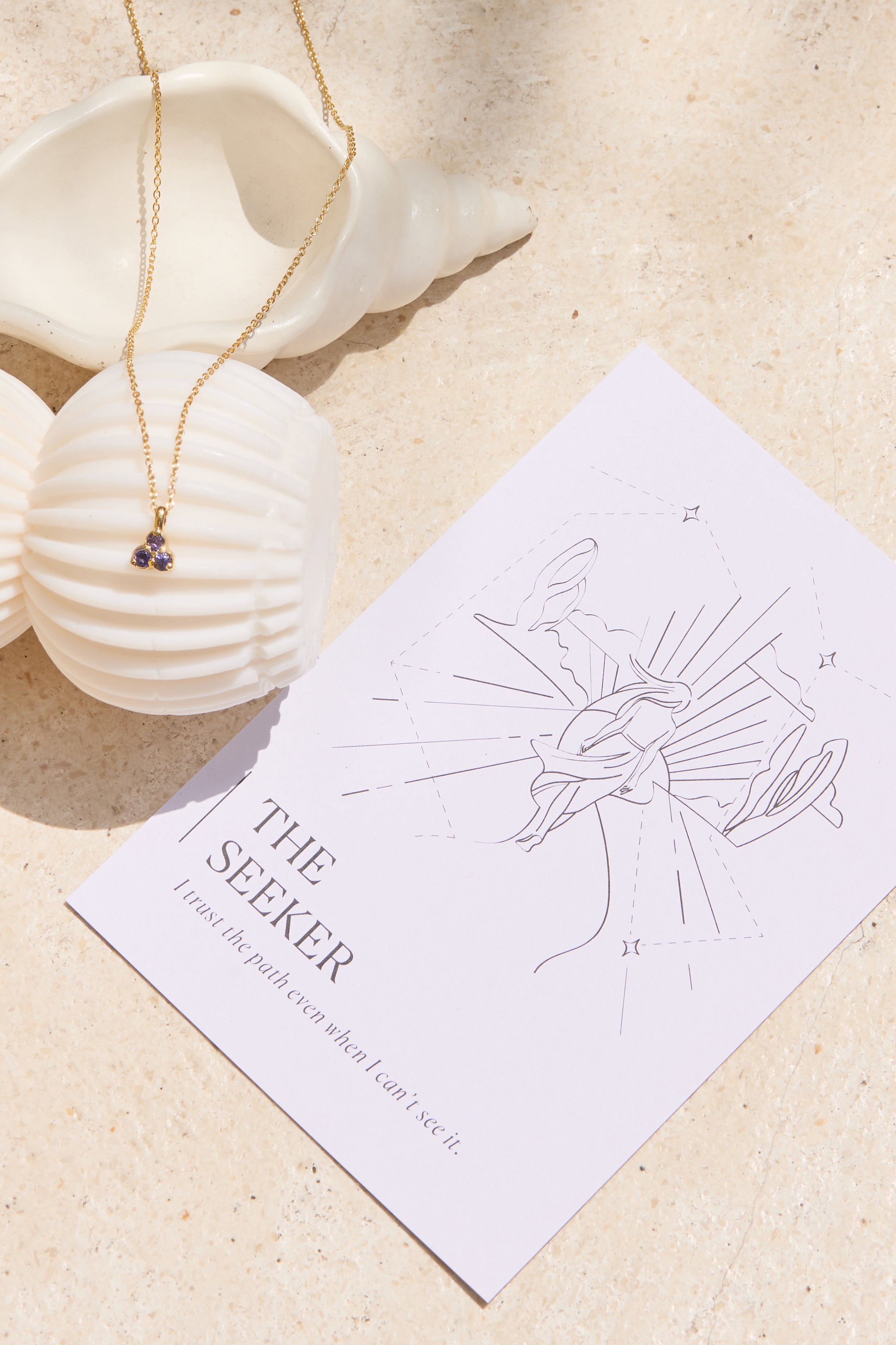 Necklace with a small triangle blue iolite pendant on a affirmation card titled 'The Seeker' on a sandy background
