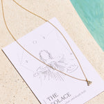 Gold necklace with aquamarine triangle pendant on a affirmation card with 'The Solace' text on a sandy background
