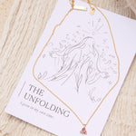 Gold necklace with a pink tourmaline triangle pendant on a affirmation card against a beige textured background