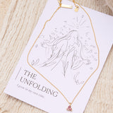 Gold necklace with a pink tourmaline triangle pendant on a affirmation card against a beige textured background