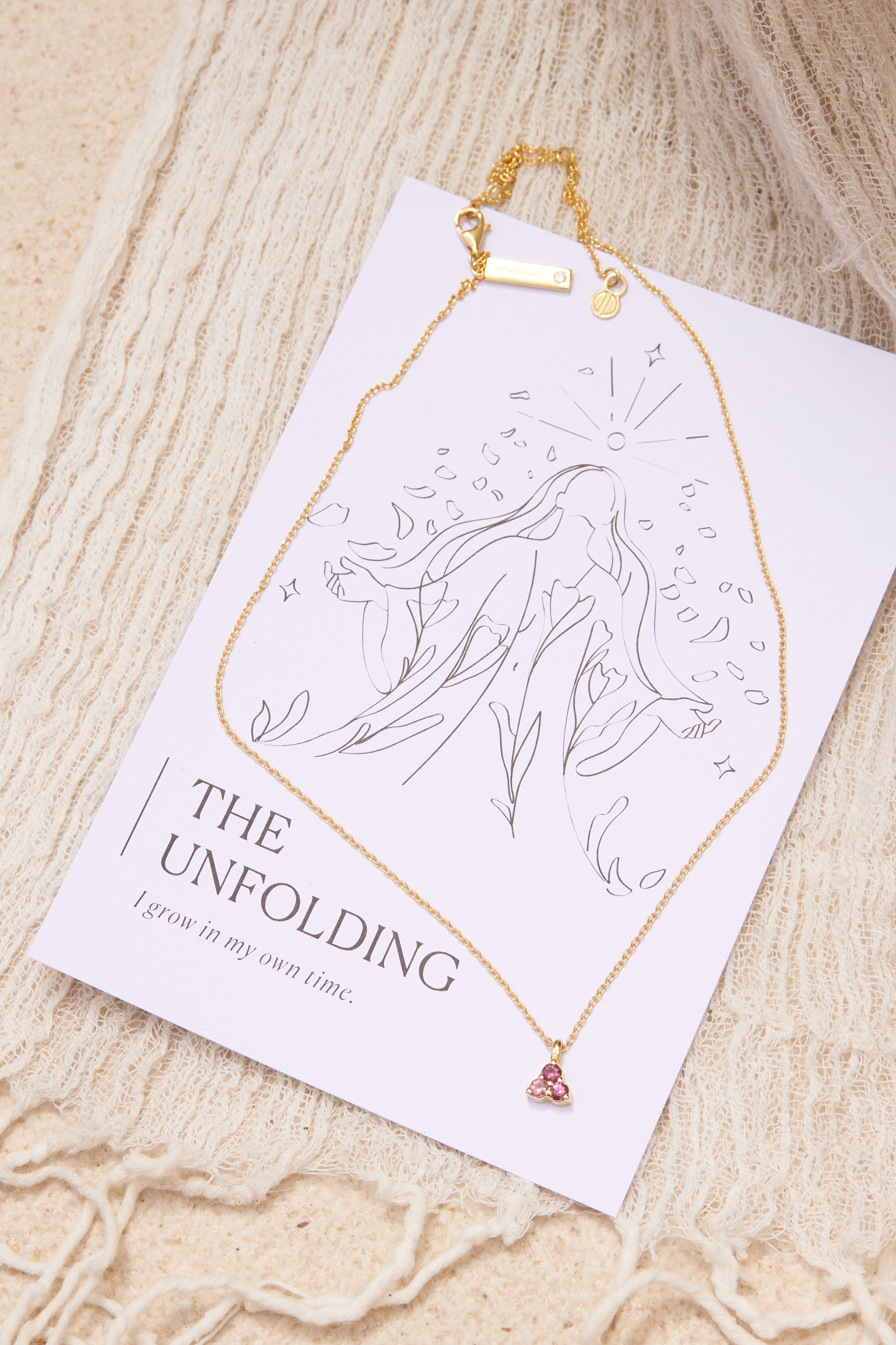 Gold necklace with a pink tourmaline triangle pendant on a affirmation card against a beige textured background